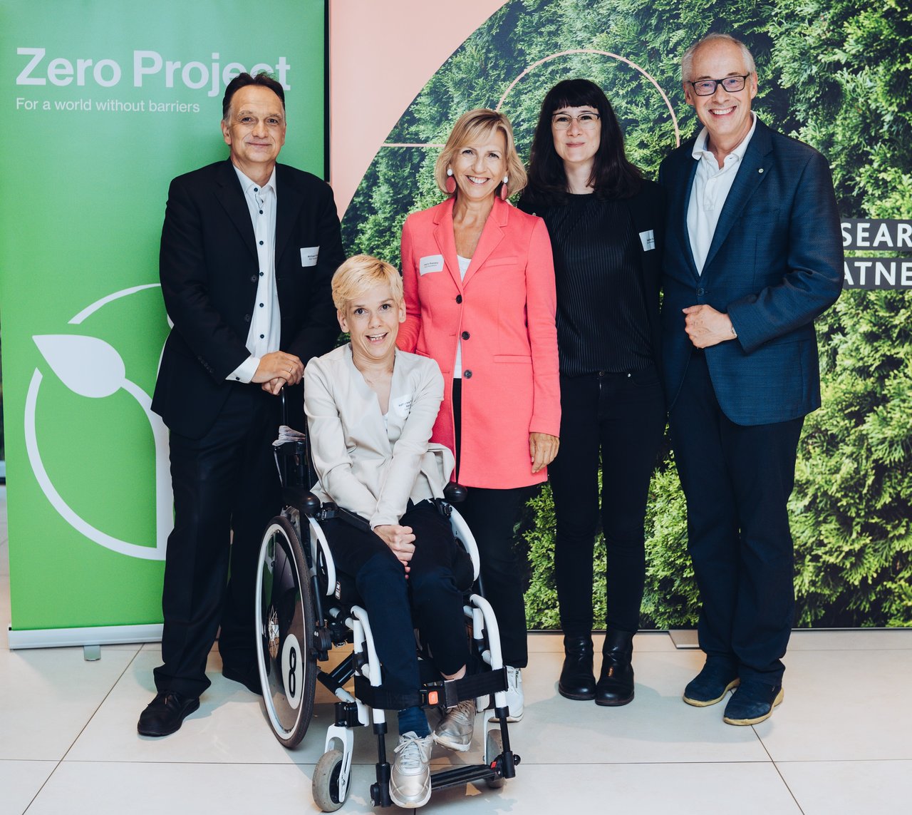 Gruppenfoto des Zero Project Austria Teams: Drei Frauen, eine davon im Rollstuhl und zwei Männer befinden sich vor dem Zero Project Rollup.